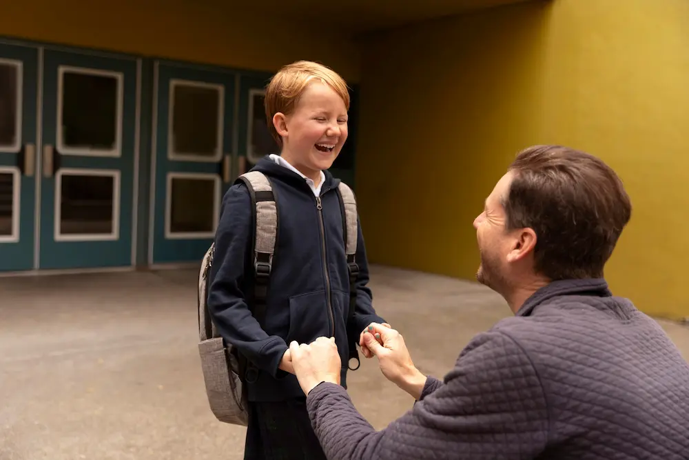 A young boy wearing a blue jacket and carrying a grey backpack beams with joy as his father crouches in front of him, holding his hands. Both are smiling and laughing, sharing a heartfelt moment as the father drops him off for his first day of school.