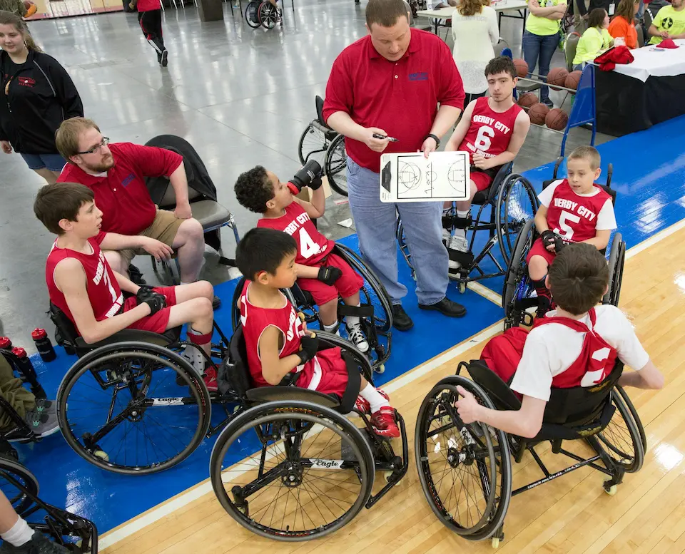A boys' wheelchair basketball team gathers around their coach on the sidelines during a break in play. The coach holds up a clipboard, illustrating a strategy as the players attentively study the plan.