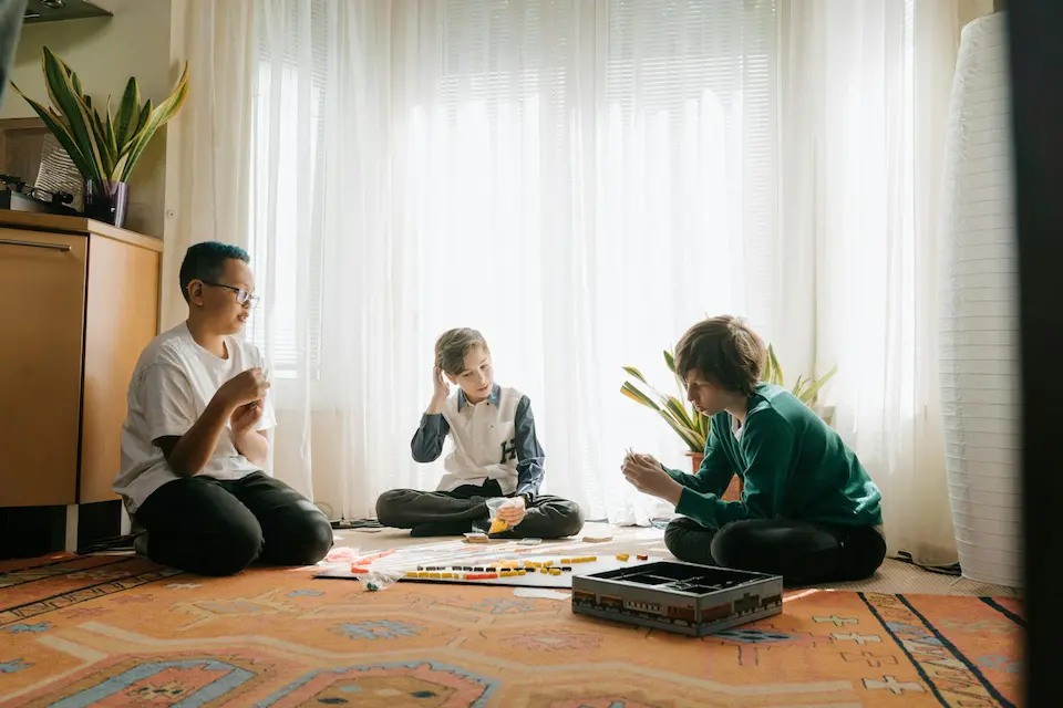 Three boys sit cross-legged on a carpeted floor in front of a draped window, deeply focused on a game of Ticket to Ride: Europe, a strategic board game about railway routes.