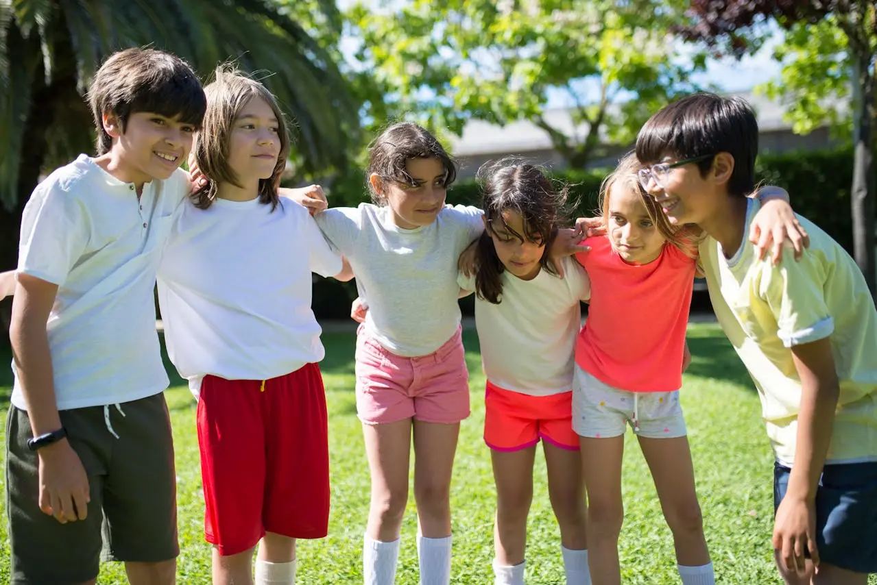 A group of children stand in a semi-circle on a field of green grass, arms wrapped around each other's shoulders, symbolizing camaraderie and togetherness.