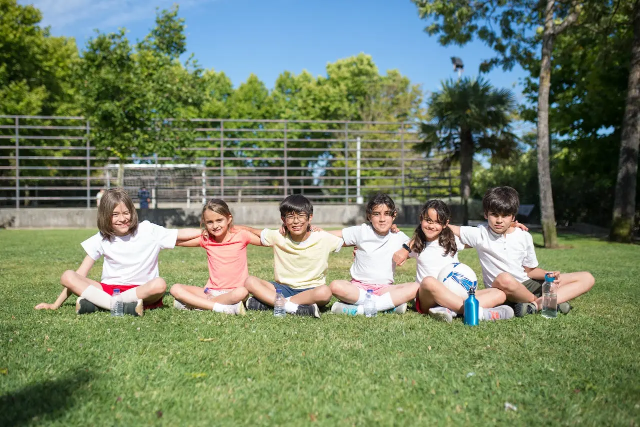 A group of children sit cross-legged on the grass in a line, arms wrapped around each other's shoulders. Their expressions reflect warmth, friendship, and togetherness in an outdoor setting.