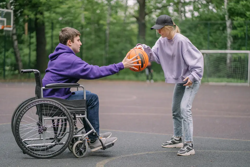 A man seated in a wheelchair and a woman stand on an outdoor basketball court, both placing one hand on a basketball, symbolizing shared participation and adaptive sports.